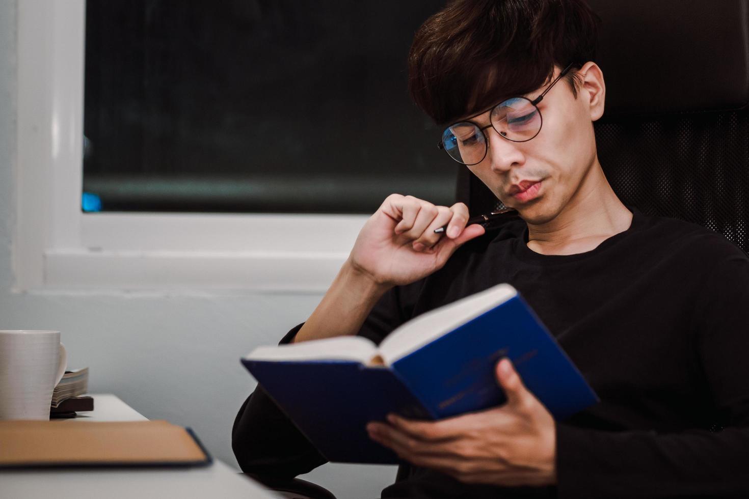 Joven apuesto hombre asiático leyendo un libro en la mesa de trabajo a altas horas de la noche foto