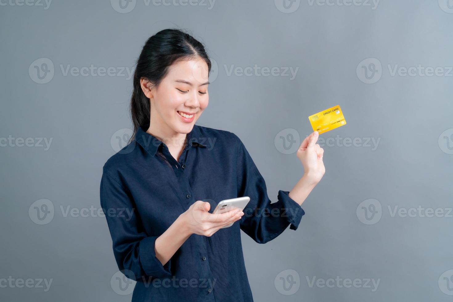 Retrato de una joven asiática feliz mostrando una tarjeta de crédito de plástico mientras sostiene el teléfono móvil sobre fondo gris foto