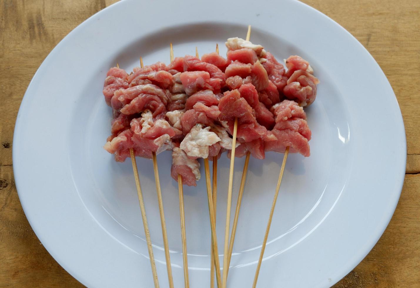 Satay de cabra cruda sobre placa blanca con mesa de madera foto