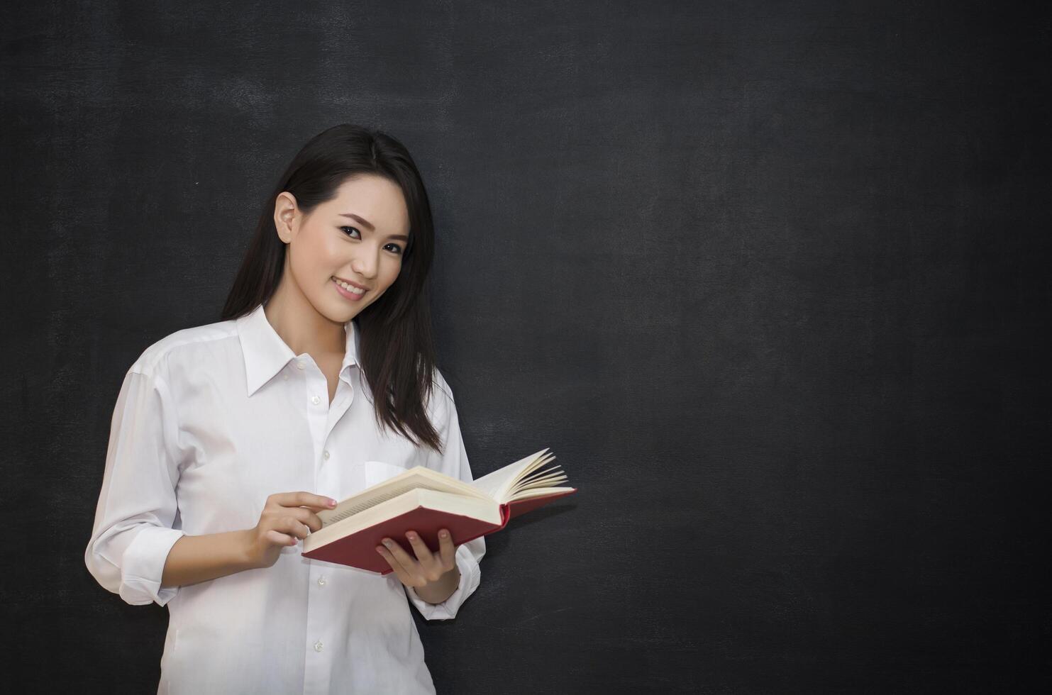 young asian woman reading book while standing against blackboard photo