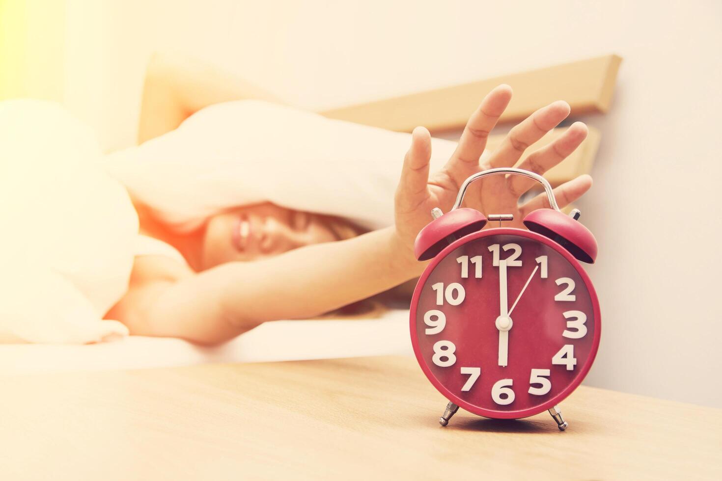 Young woman frowning in bed and holding hand on red alarm clock. photo