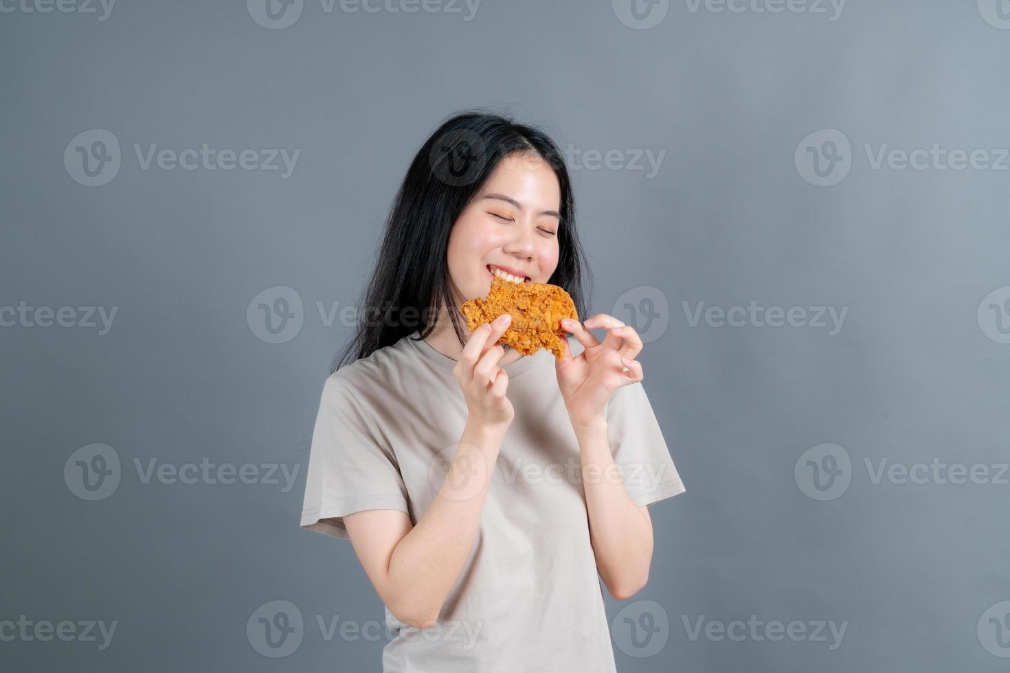 Young Asian woman wearing t-shirt with happy face and enjoy eating fried chicken on grey background photo