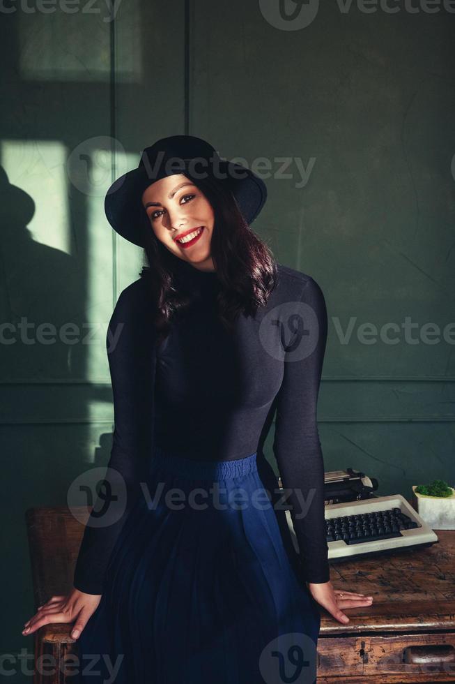 Hermosa mujer con sombrero se sienta cerca de la mesa de roble con máquina de escribir vintage foto