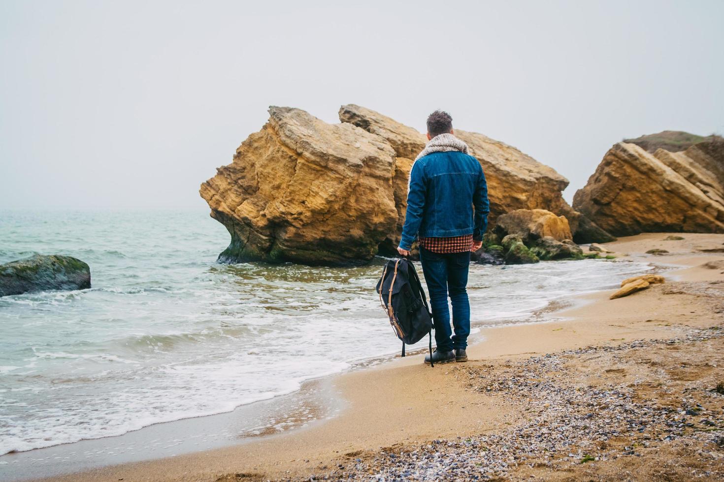 Hombre con una mochila de pie cerca de una roca frente a un hermoso mar foto