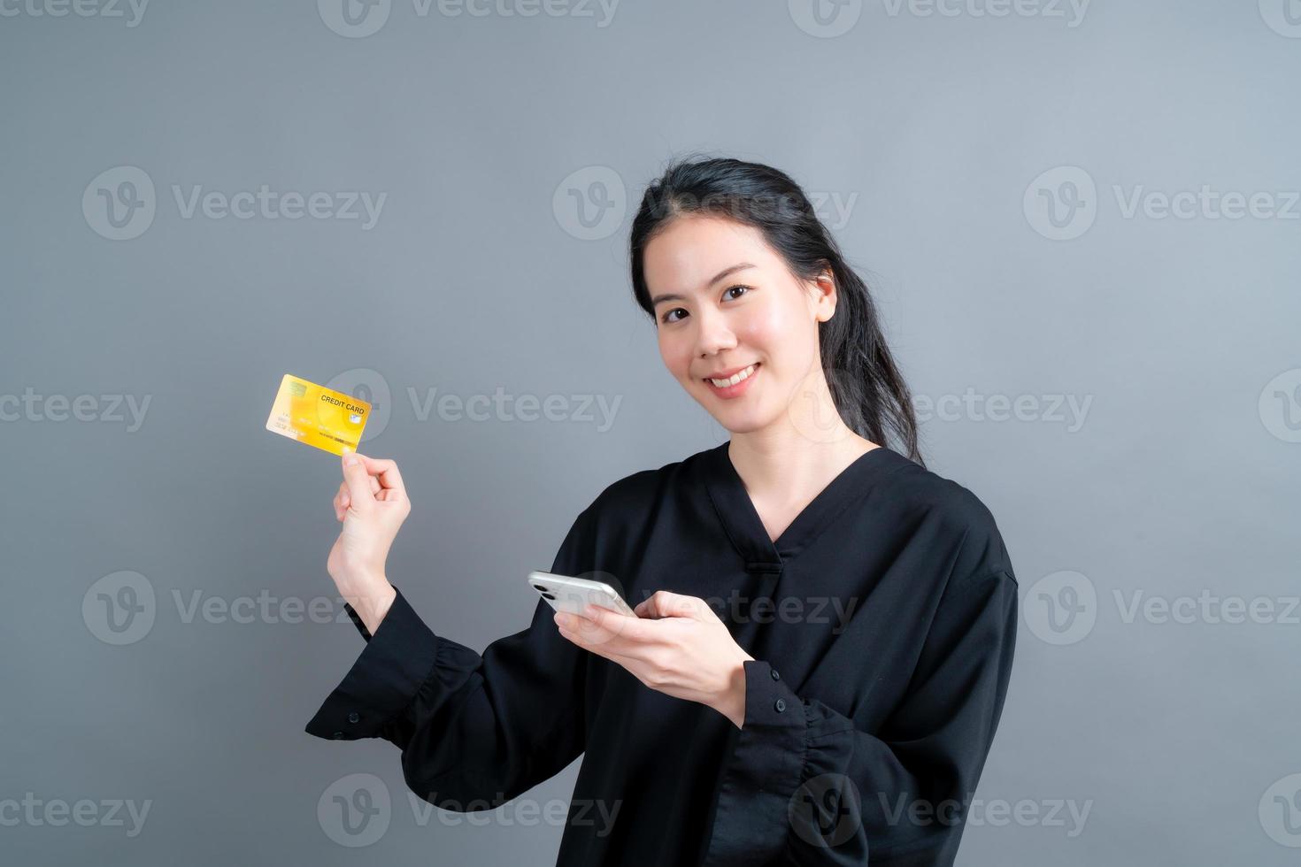 Retrato de una joven asiática feliz mostrando una tarjeta de crédito de plástico mientras sostiene el teléfono móvil sobre fondo gris foto