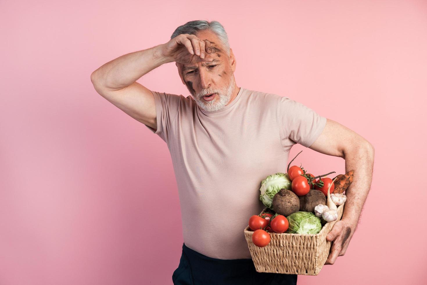 Tired senior man holding a basket with vegetables on a pink background photo