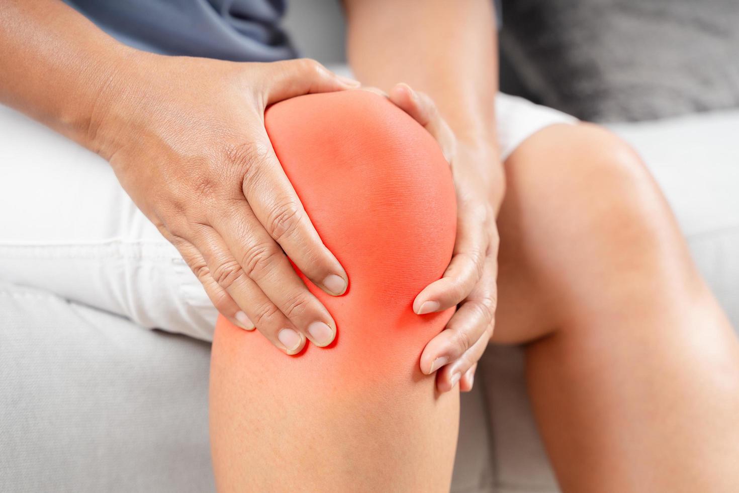 Woman sitting on sofa feeling knee pain and massages her knee photo