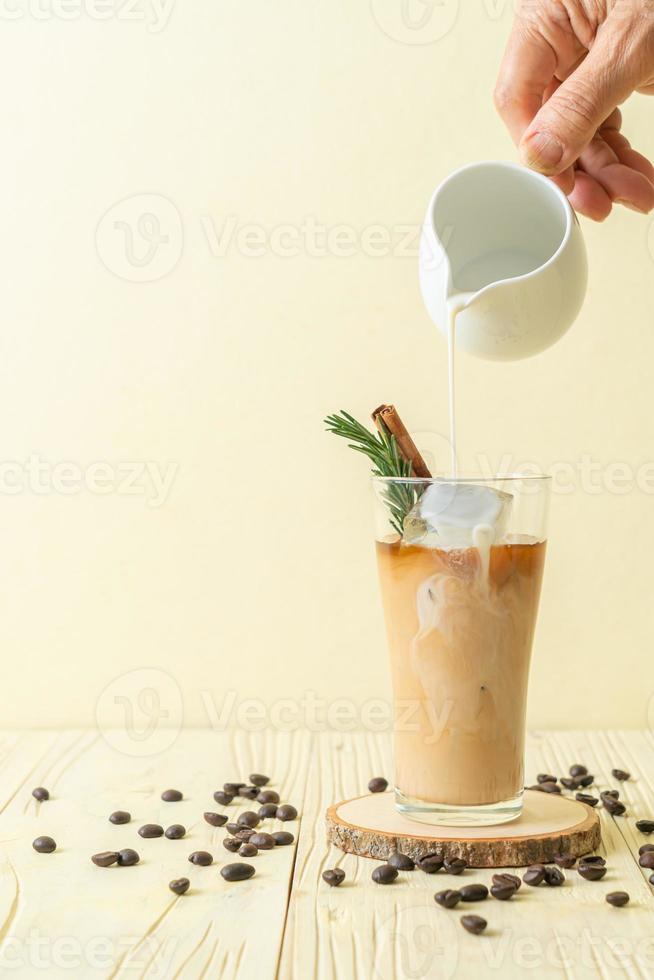 Pouring milk in black coffee glass with ice cube, cinnamon, and rosemary on wood background photo