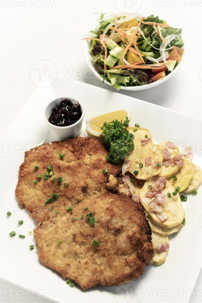 German breaded organic pork schnitzel with bacon fried potatoes cranberry sauce and salad on white studio background photo