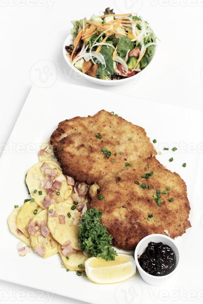 German breaded organic pork schnitzel with bacon fried potatoes cranberry sauce and salad on white studio background photo