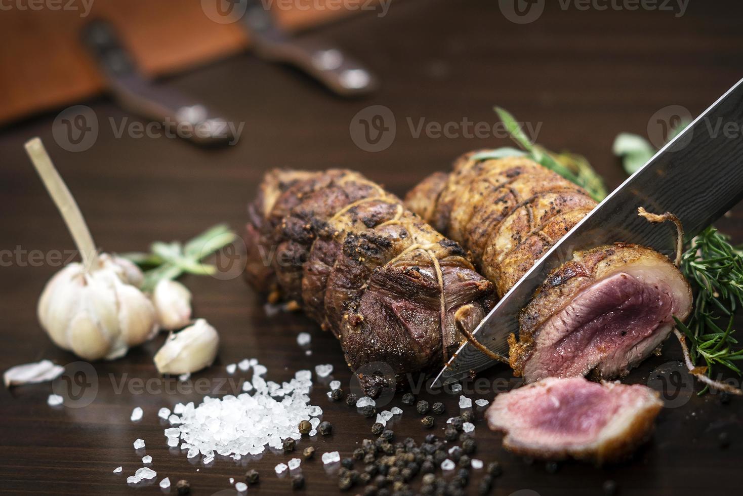 Cortar una porción de rosbif orgánico con un cuchillo en la mesa de madera con ajo, pimienta y sal en Melbourne, Australia foto