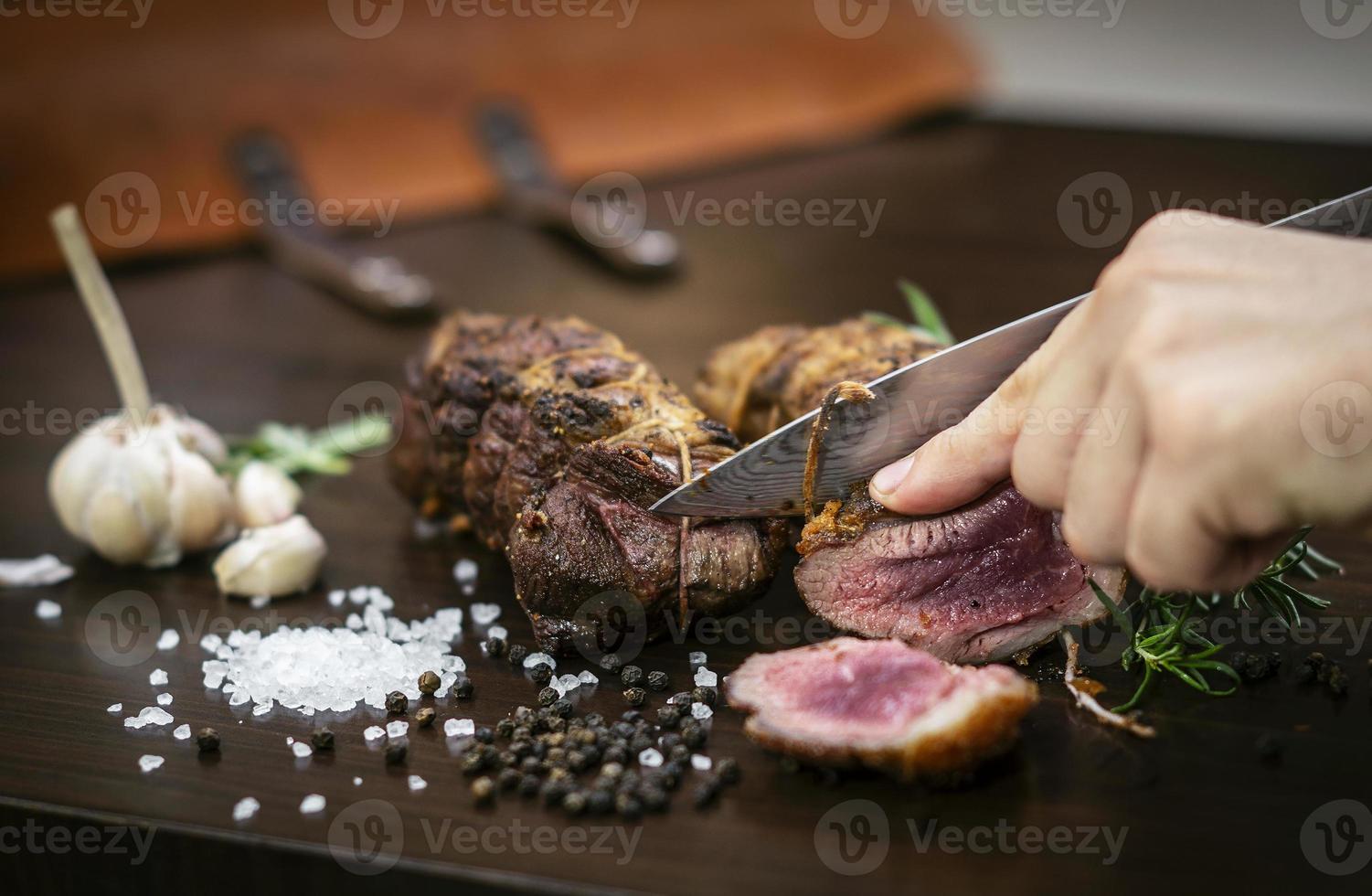 Cortar una porción de rosbif orgánico con un cuchillo en la mesa de madera con ajo, pimienta y sal en Melbourne, Australia foto