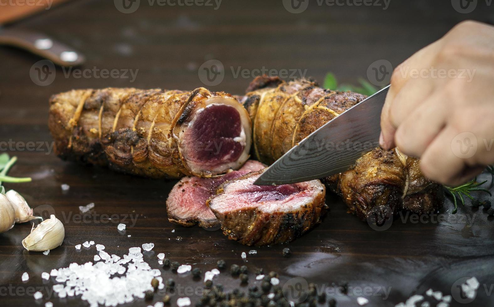 Slicing a serving of organic roast beef roll with knife on wood table with garlic pepper and salt in Melbourne Australia photo