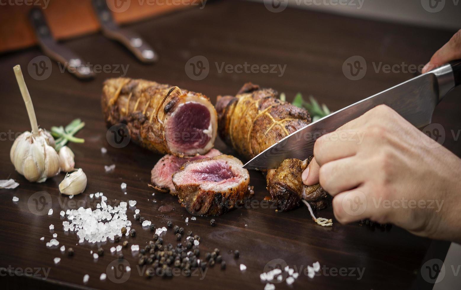 Slicing a serving of organic roast beef roll with knife on wood table with garlic pepper and salt in Melbourne Australia photo