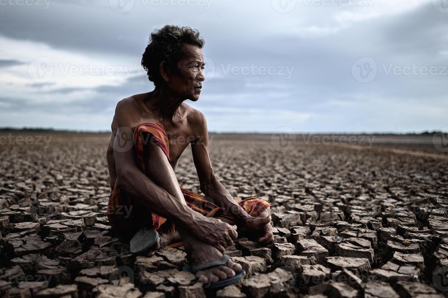 un anciano sentado abrazando sus rodillas dobladas sobre suelo seco. foto