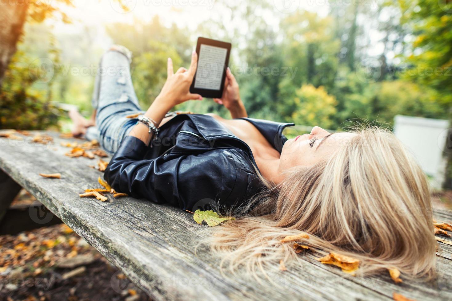 Chica rubia lee un libro electrónico acostado sobre una mesa de madera en el parque de otoño foto