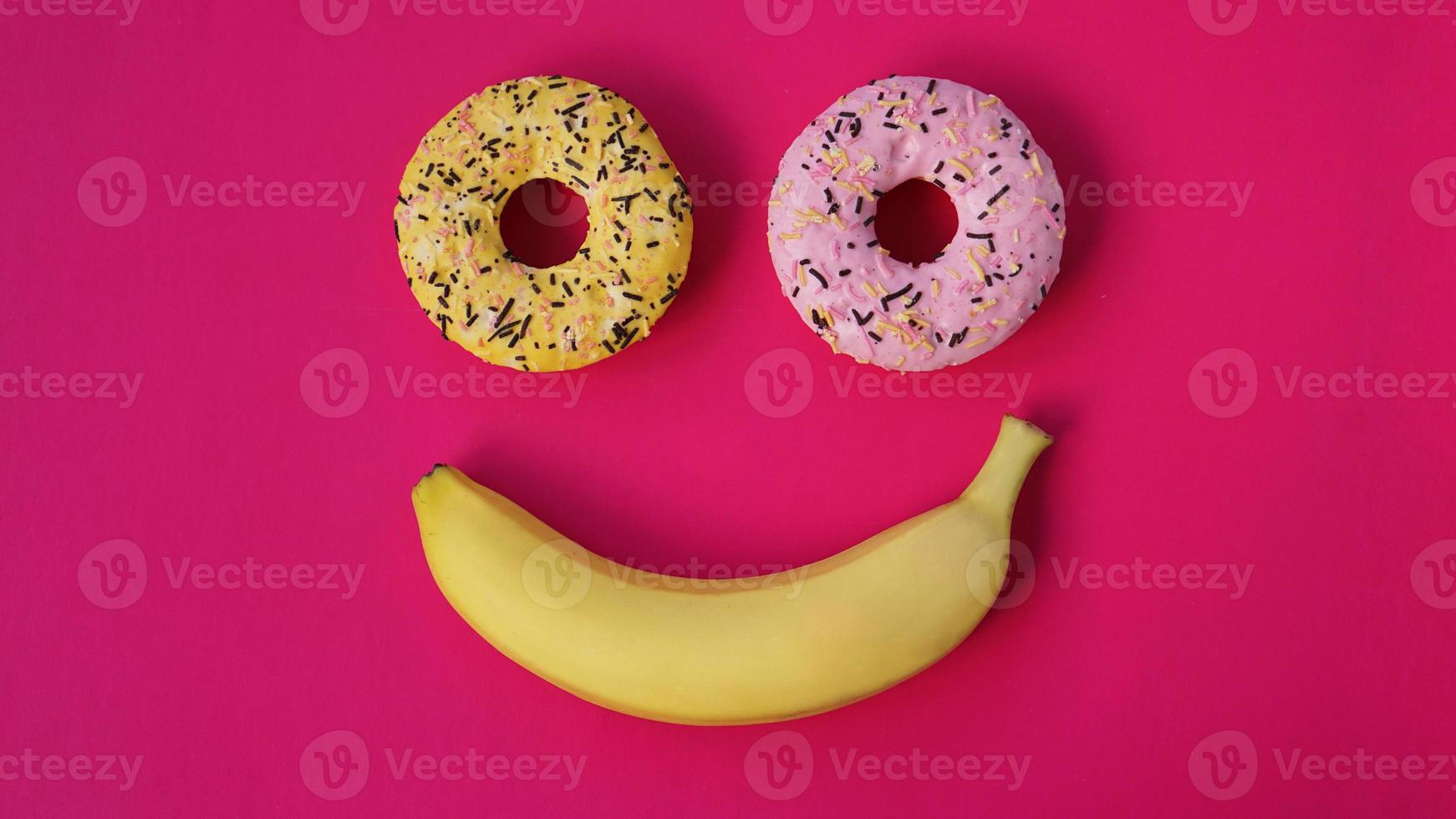 Two donuts and a banana lie on pink surface, forming a smile emotion photo