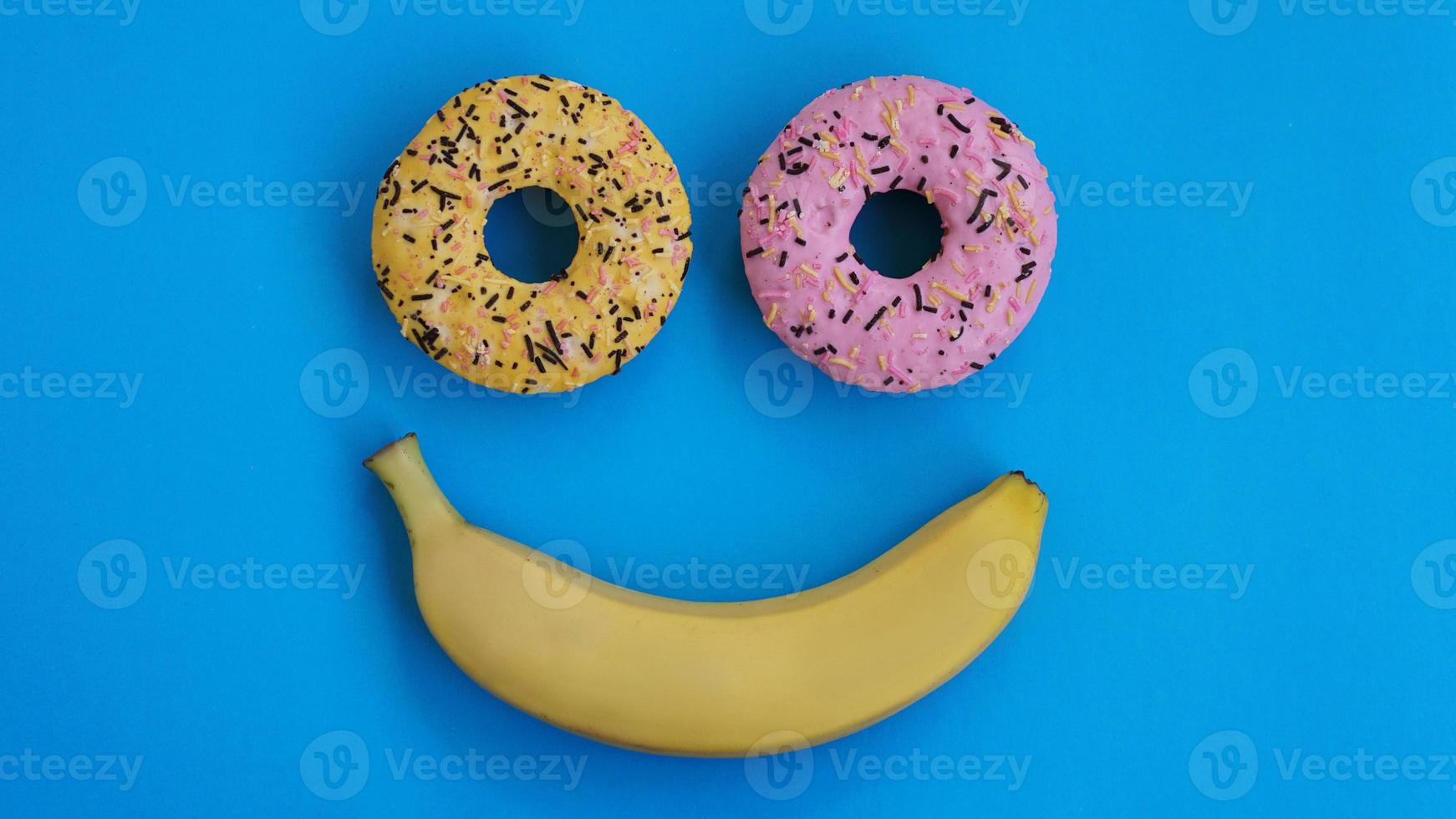 Two donuts and a banana lie on blue surface, forming a smile emotion photo