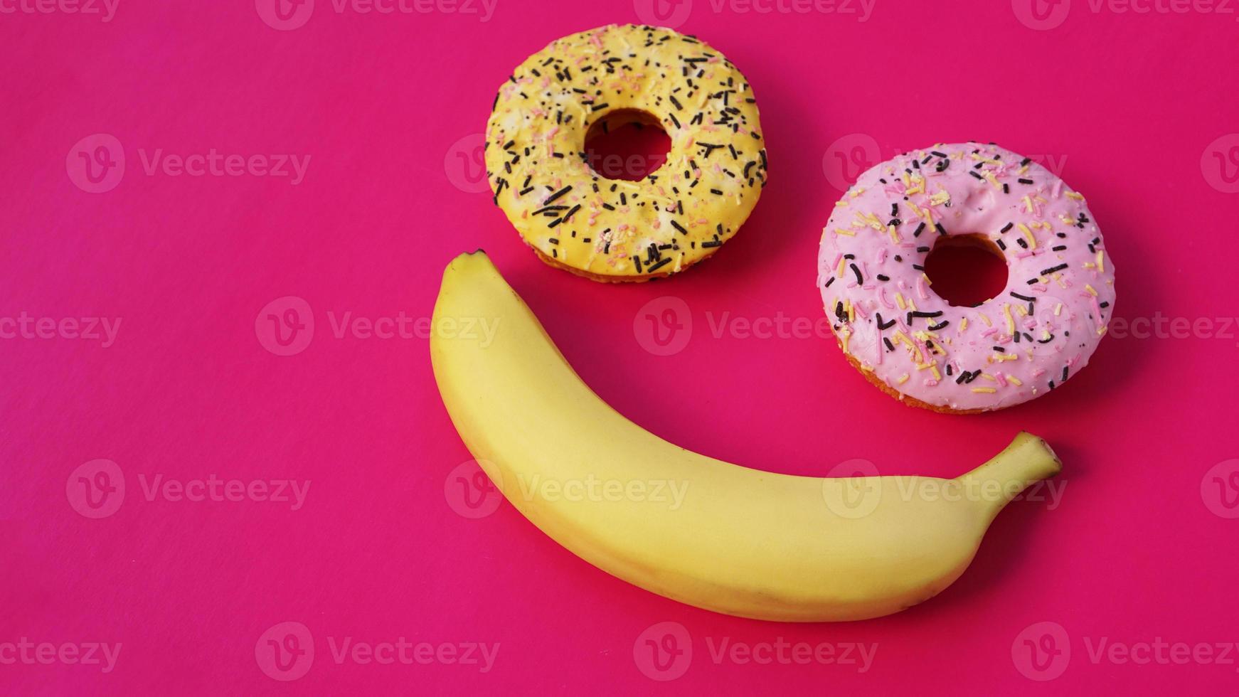 dos donas y un plátano yacen sobre una superficie rosa, formando una emoción de sonrisa foto