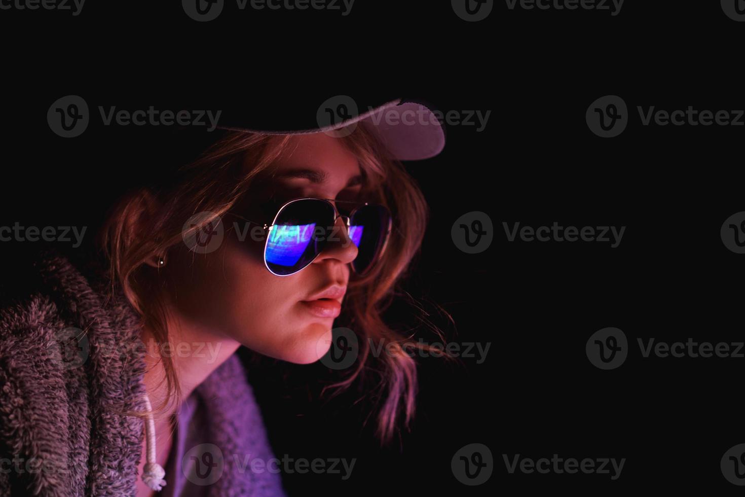 Hacker girl in cap and sunglasses in front of a computer in neon light photo