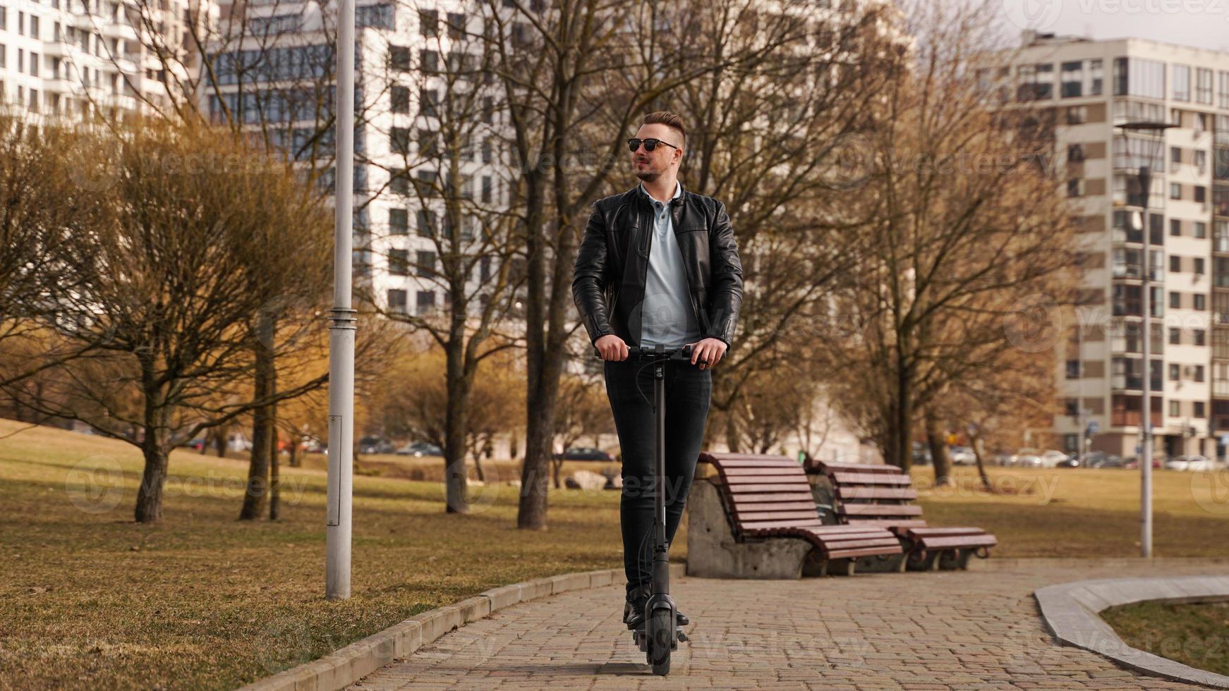 Young man in a black jacket and sunglasses rides an electronic scooter photo