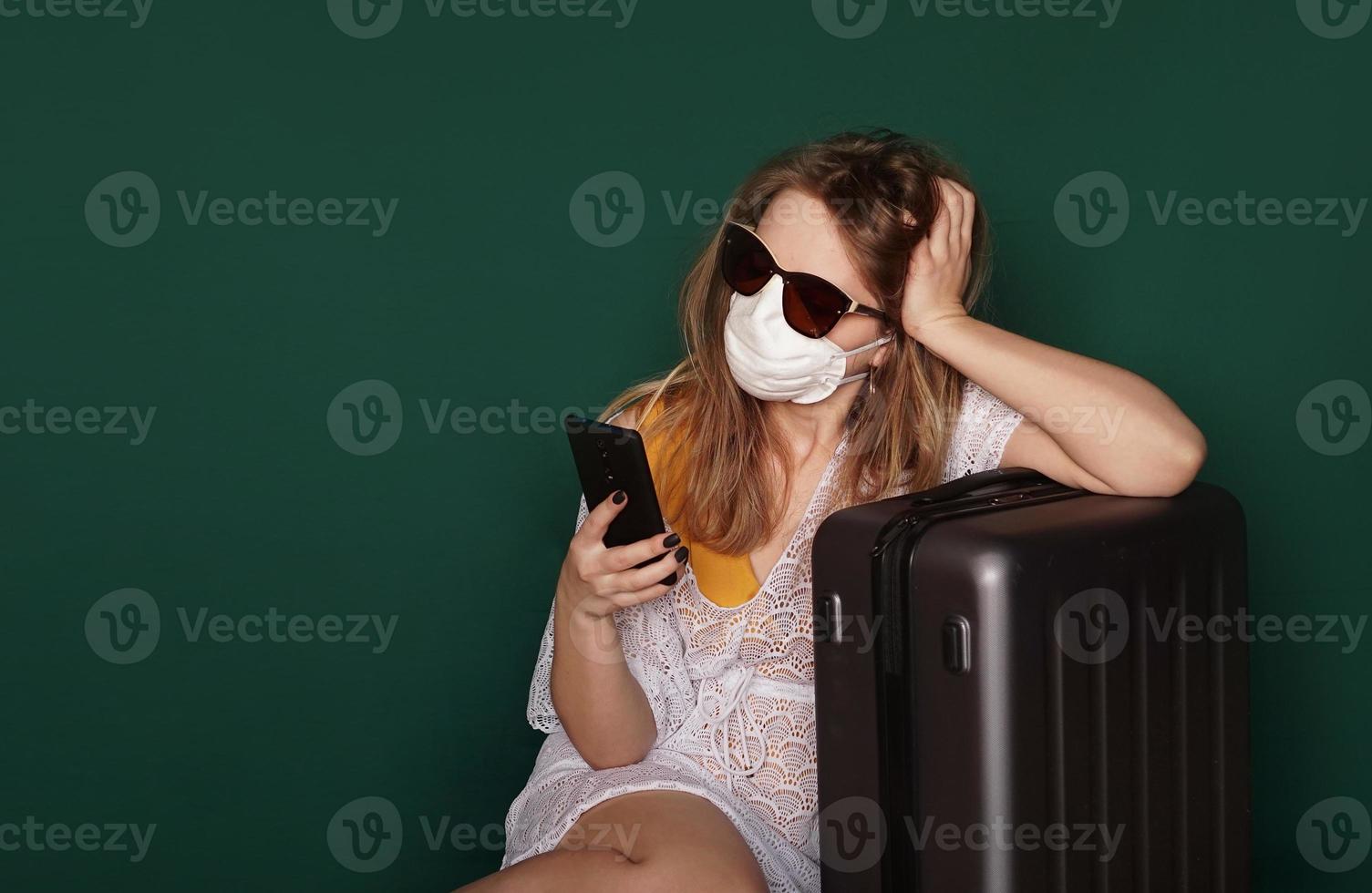 Girl tourist in a medical mask sits with luggage on a green background photo