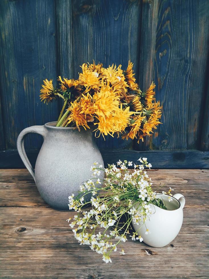 Beautiful field flowers in cup and ceramic jug on wooden backdrop photo