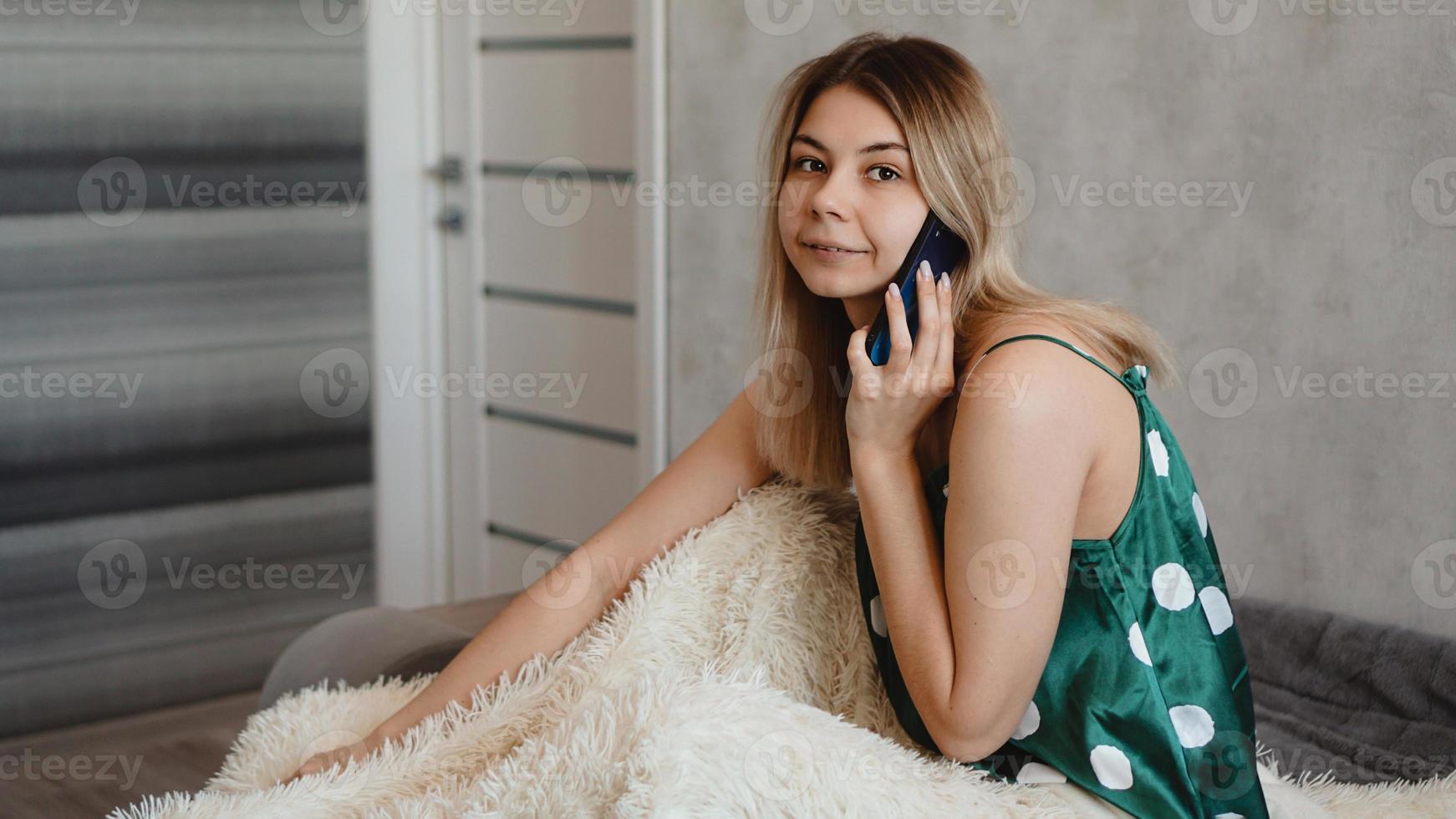 Atractiva mujer joven sentada en la cama y hablando por teléfono inteligente foto