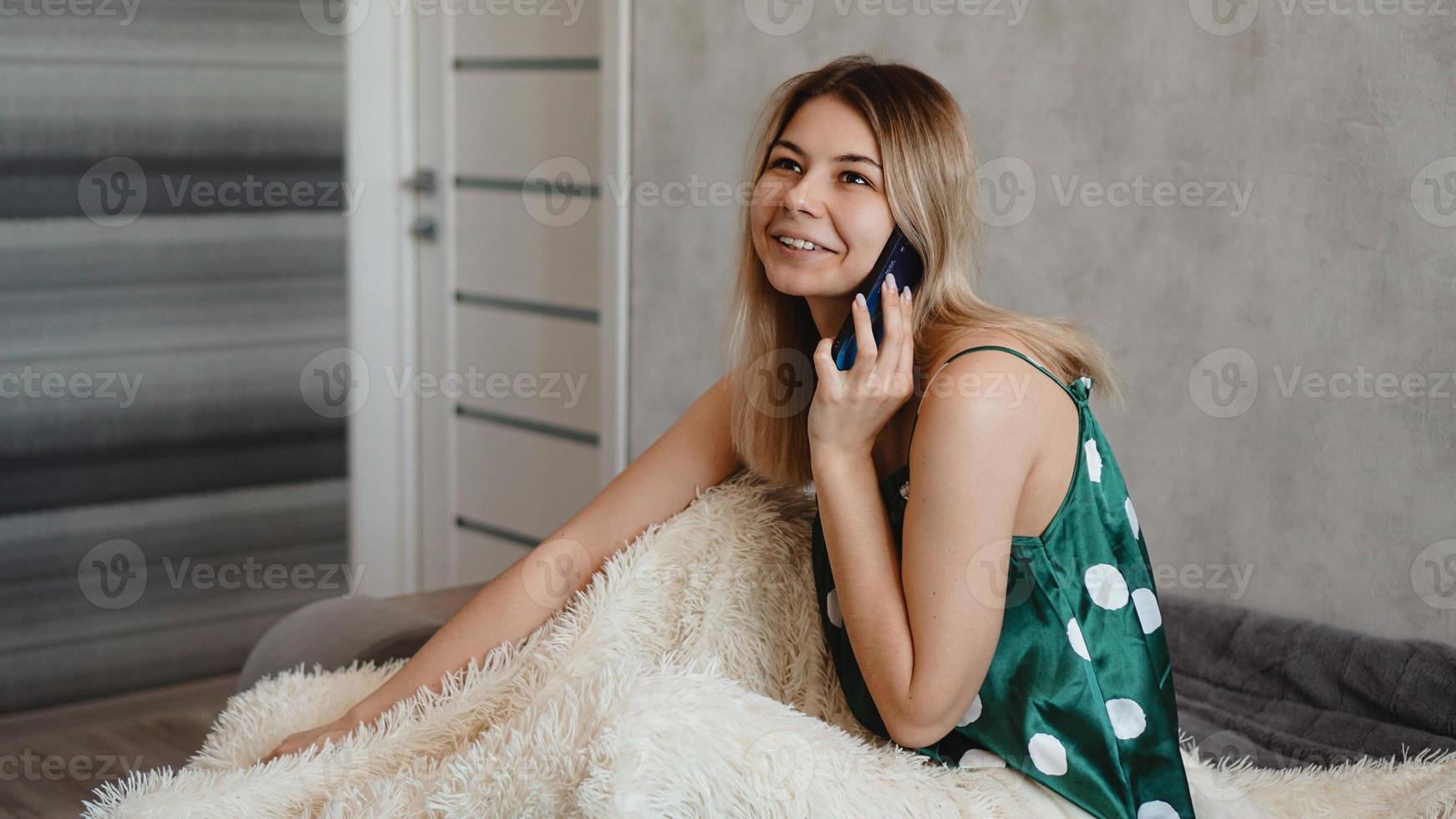 Atractiva mujer joven sentada en la cama y hablando por teléfono inteligente foto