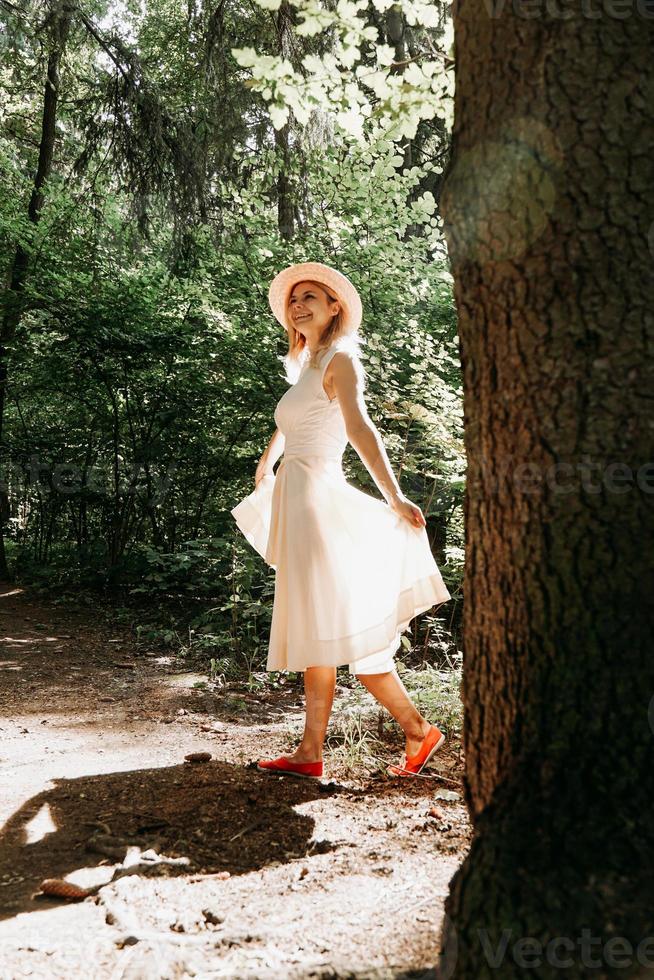A girl in a white dress and hat walks through a summer park or forest photo