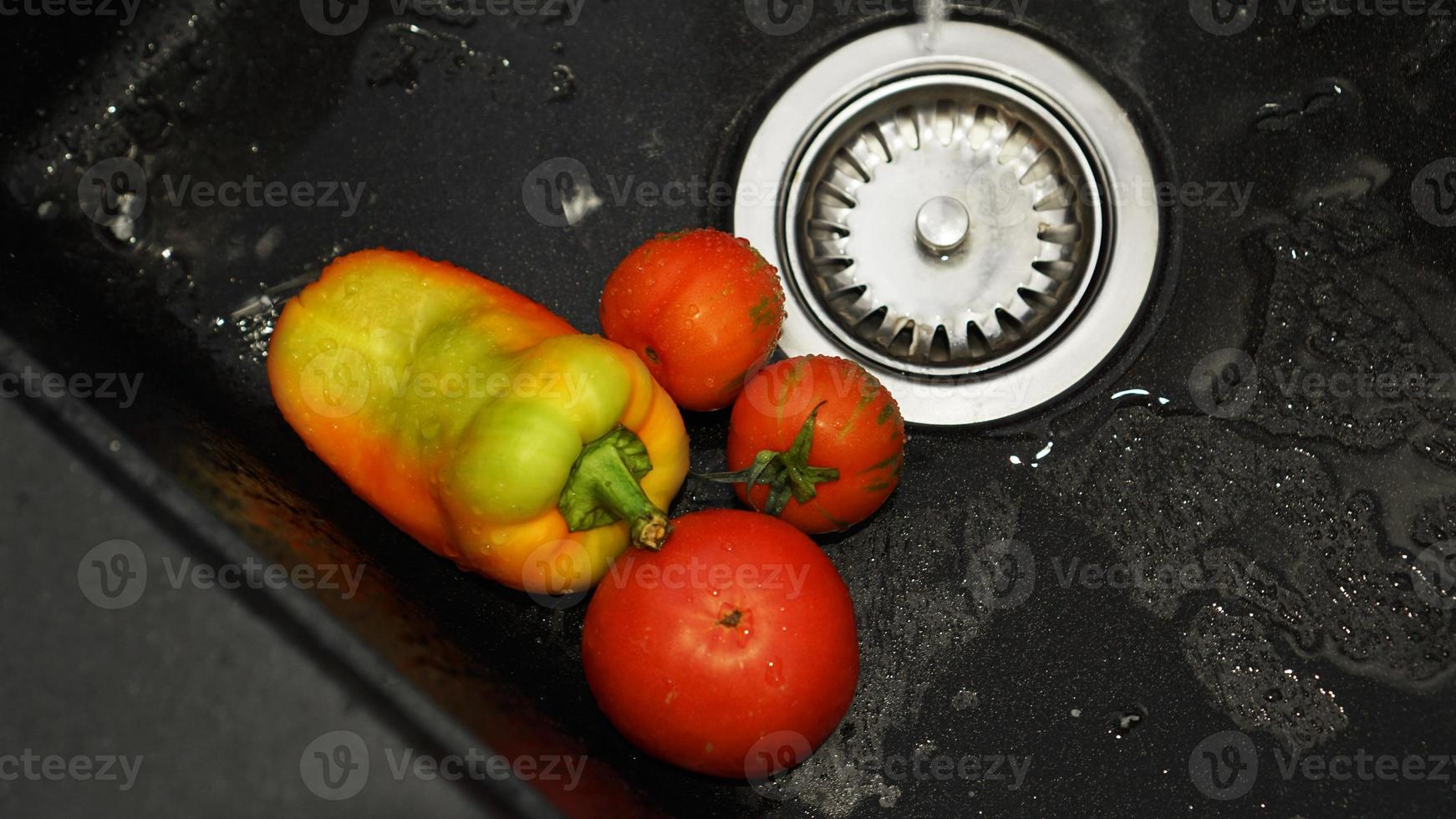Verduras: tomates y pimientos en un fregadero de cocina negro bajo el agua foto