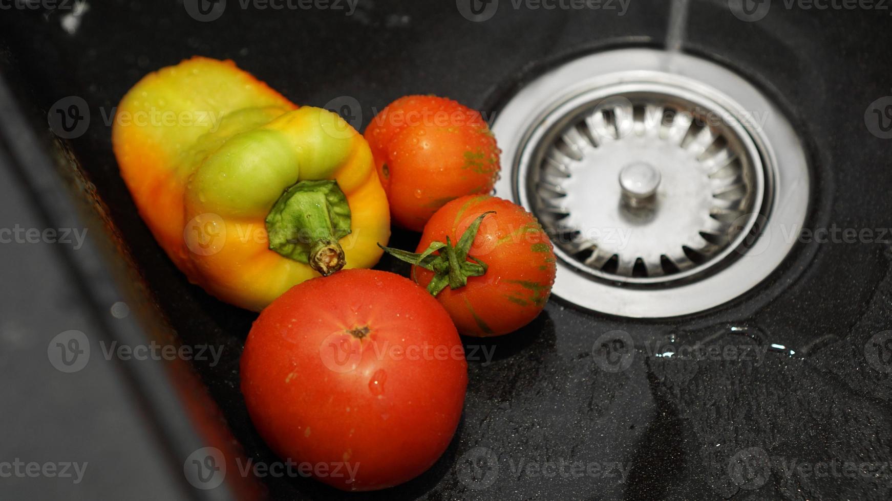 Verduras: tomates y pimientos en un fregadero de cocina negro bajo el agua foto