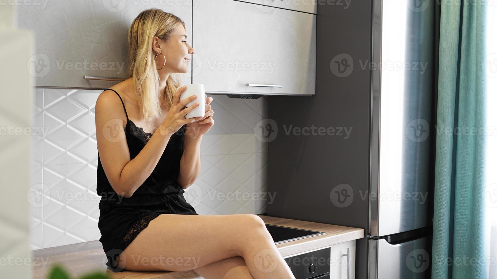 Cheerful woman sits on countertop in white modern kitchen. photo