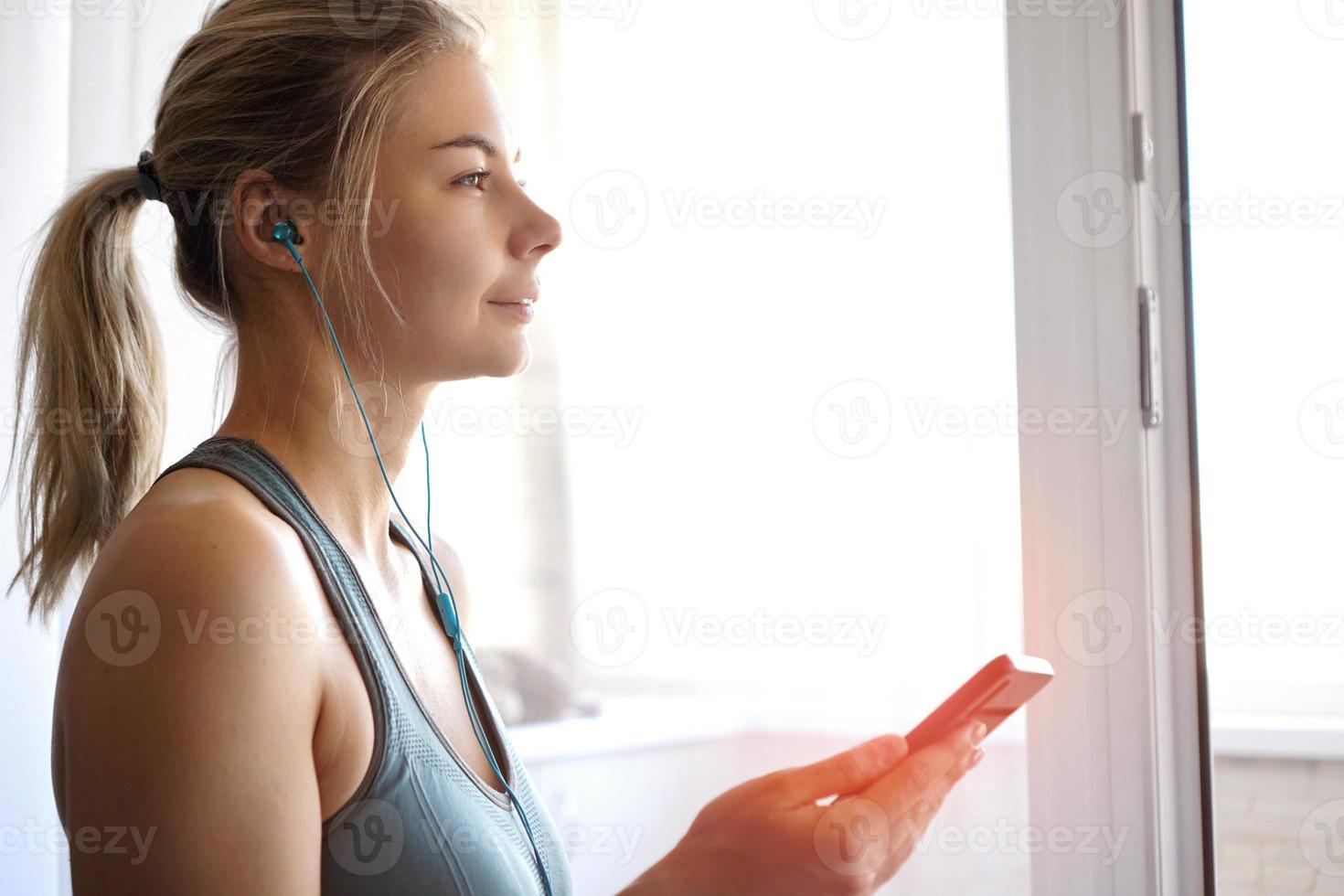 Young pretty sporty girl stands near window and listens to music photo