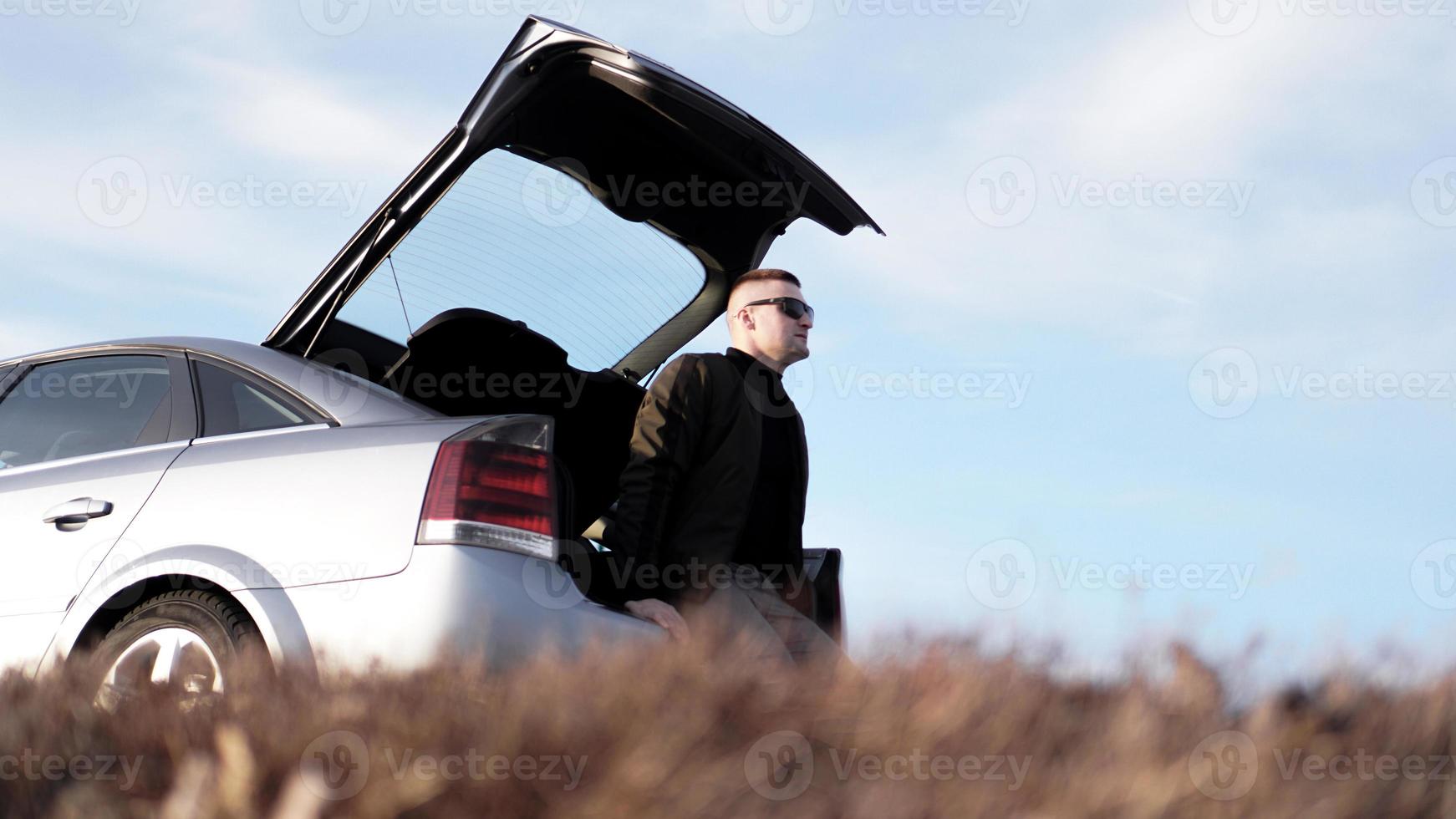 Side view of businessman in eyeglasses sitting on car trunk photo