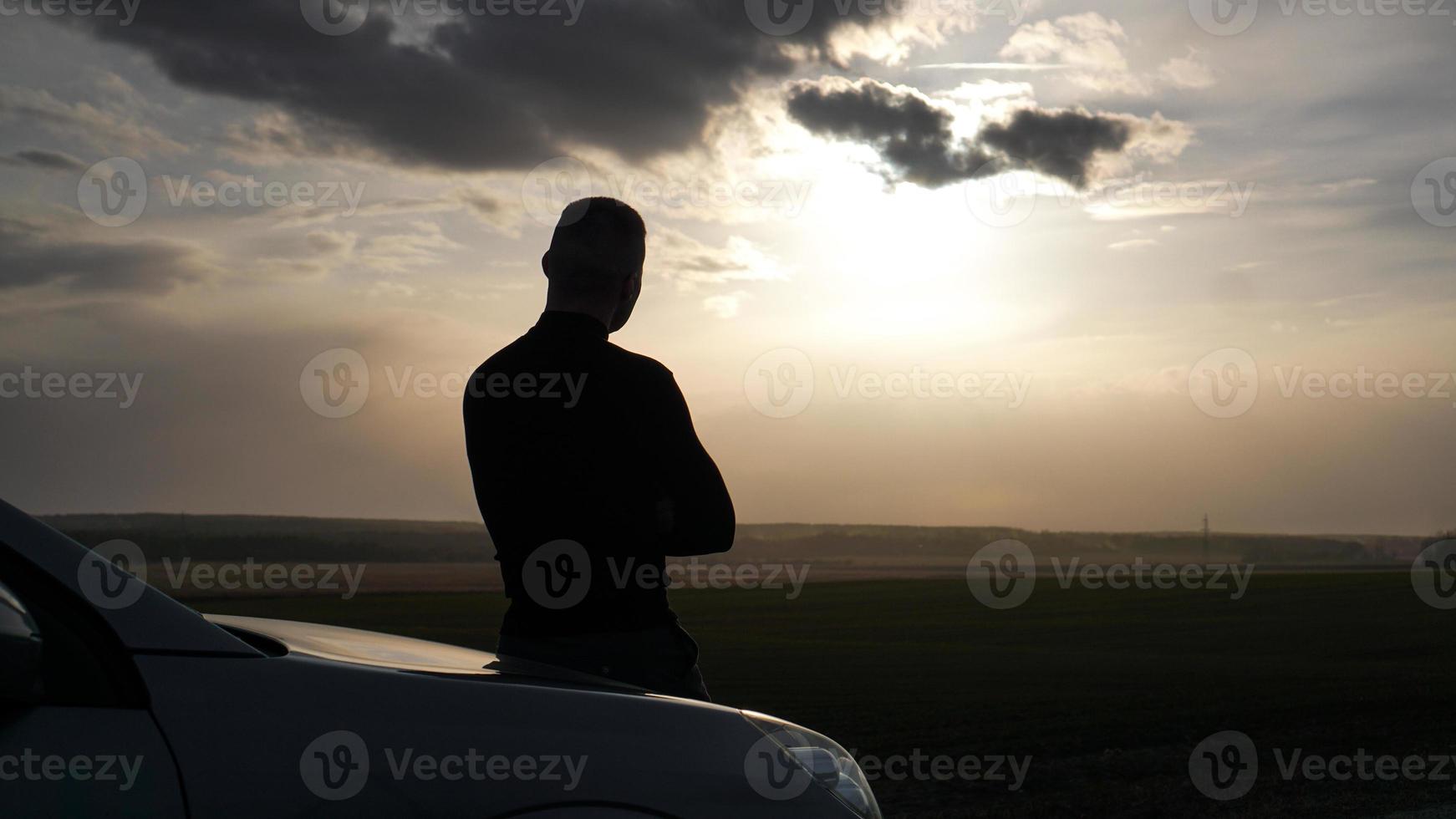 Silhouette of the man standing near to car and looking at a sunset photo