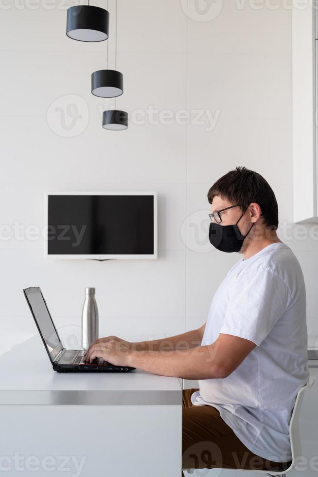 Young man in black protective mask working with laptop from home photo