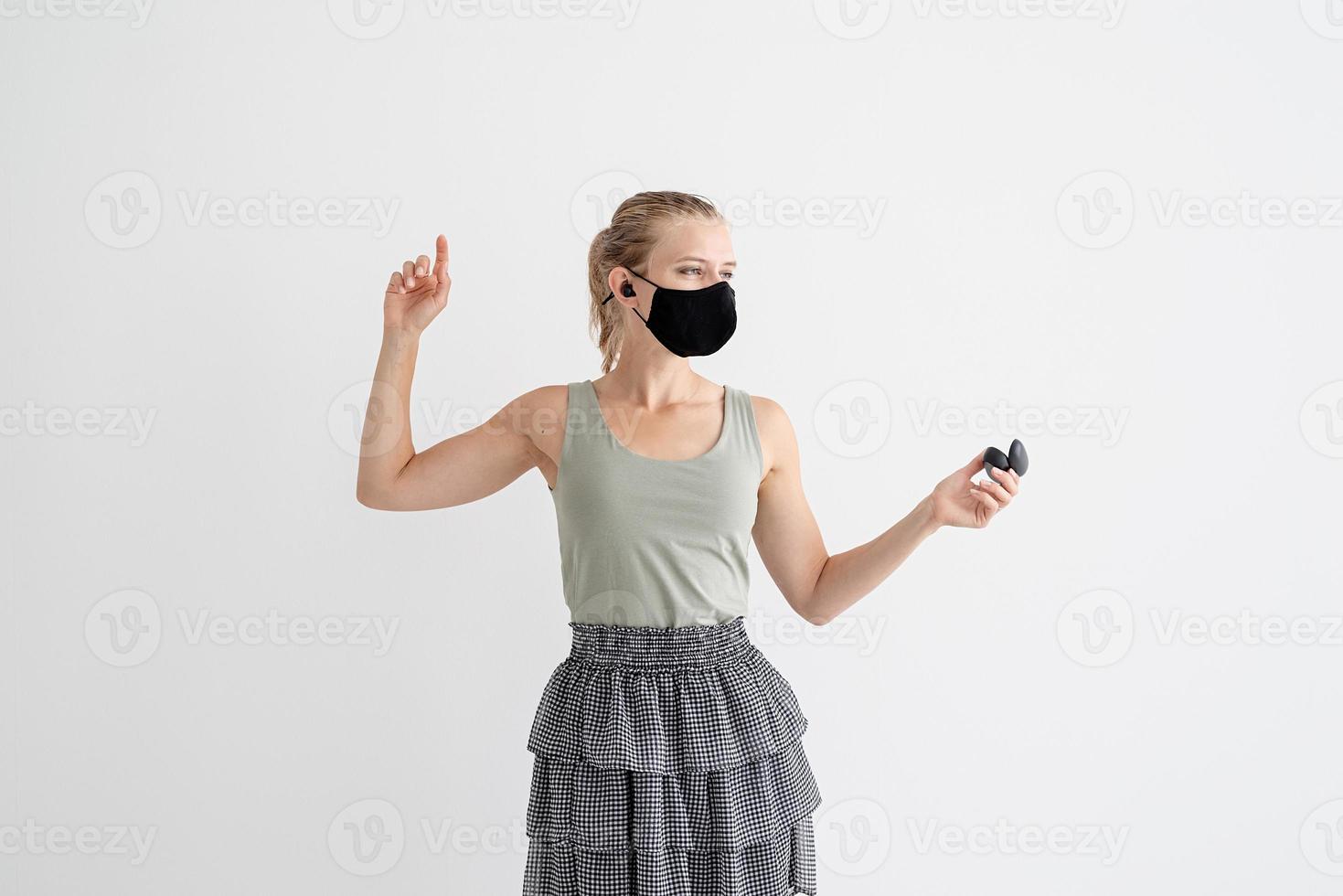 Young woman in a protective mask holding wireless earbuds and dancing photo