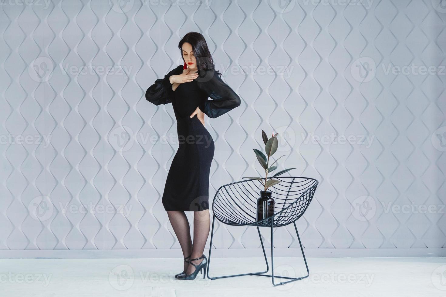 Fashion brunette woman wearing a black dress standing near chair photo