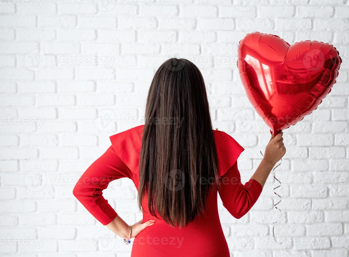 Joven mujer morena en vestido rojo sosteniendo un globo de corazón rojo foto