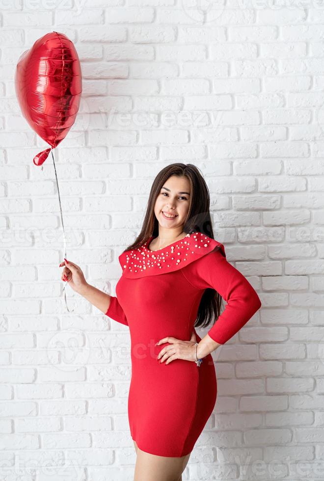 Joven mujer morena en vestido rojo sosteniendo un globo de corazón rojo foto