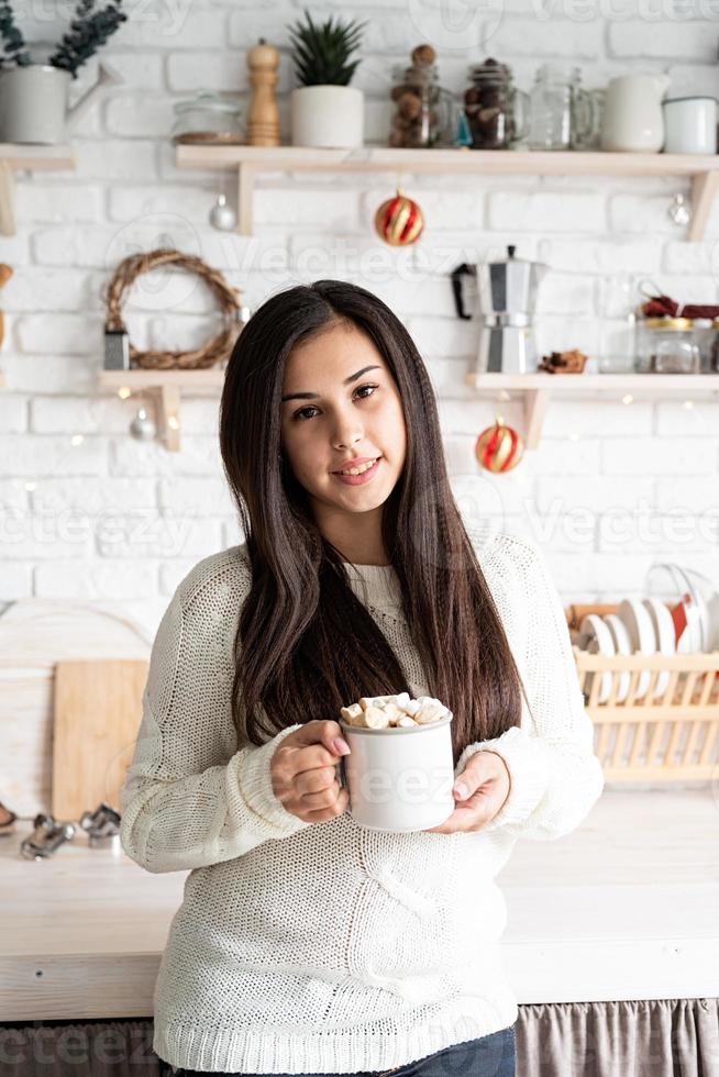 Mujer morena sosteniendo una taza de chocolate con malvaviscos en la cocina foto