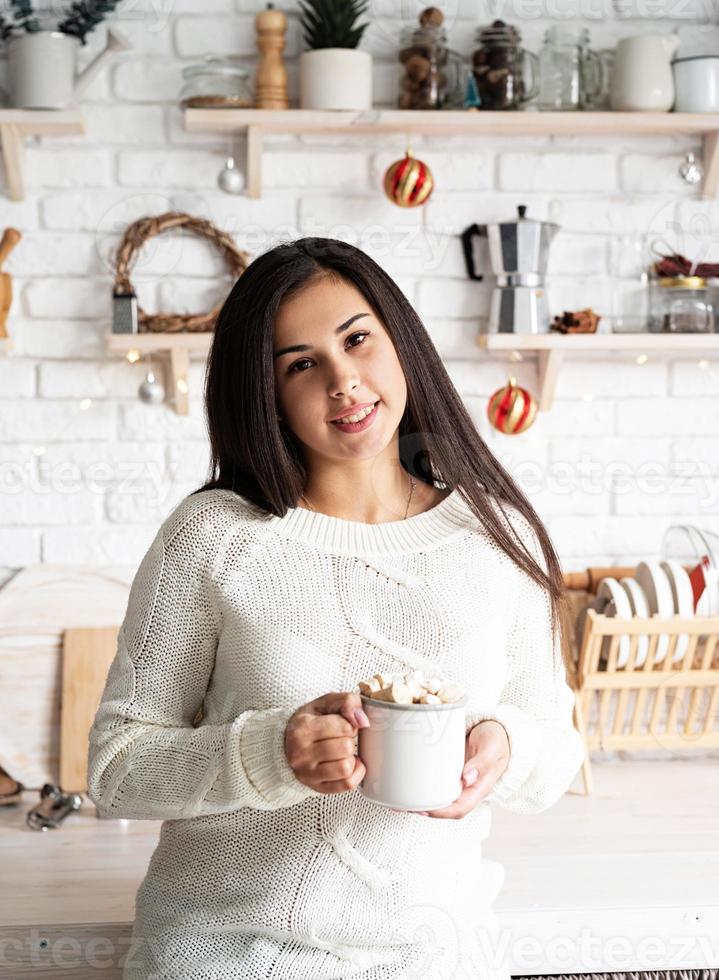 Mujer morena sosteniendo una taza de chocolate con malvaviscos en la cocina foto