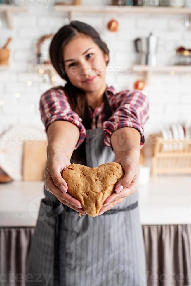 Happy brunette woman holding heart shaped dough at the kitchen photo