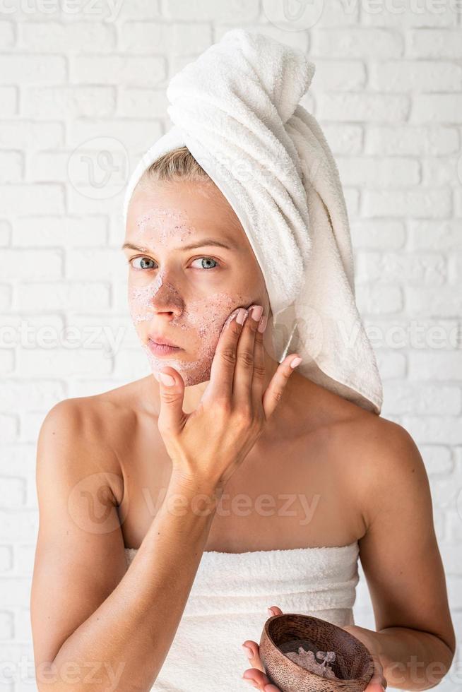 Mujer preocupada en toallas de baño blancas aplicando exfoliante en su rostro foto