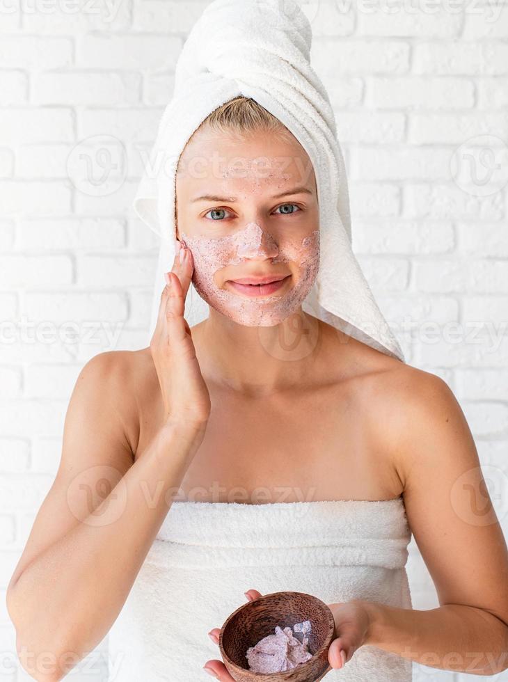 Joven mujer caucásica vistiendo toallas blancas aplicando exfoliante en su rostro foto