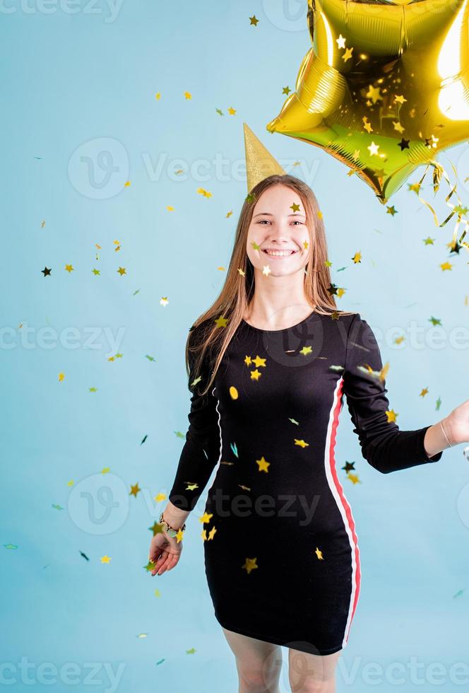 Chica adolescente soplando confeti sobre fondo azul sosteniendo globos foto