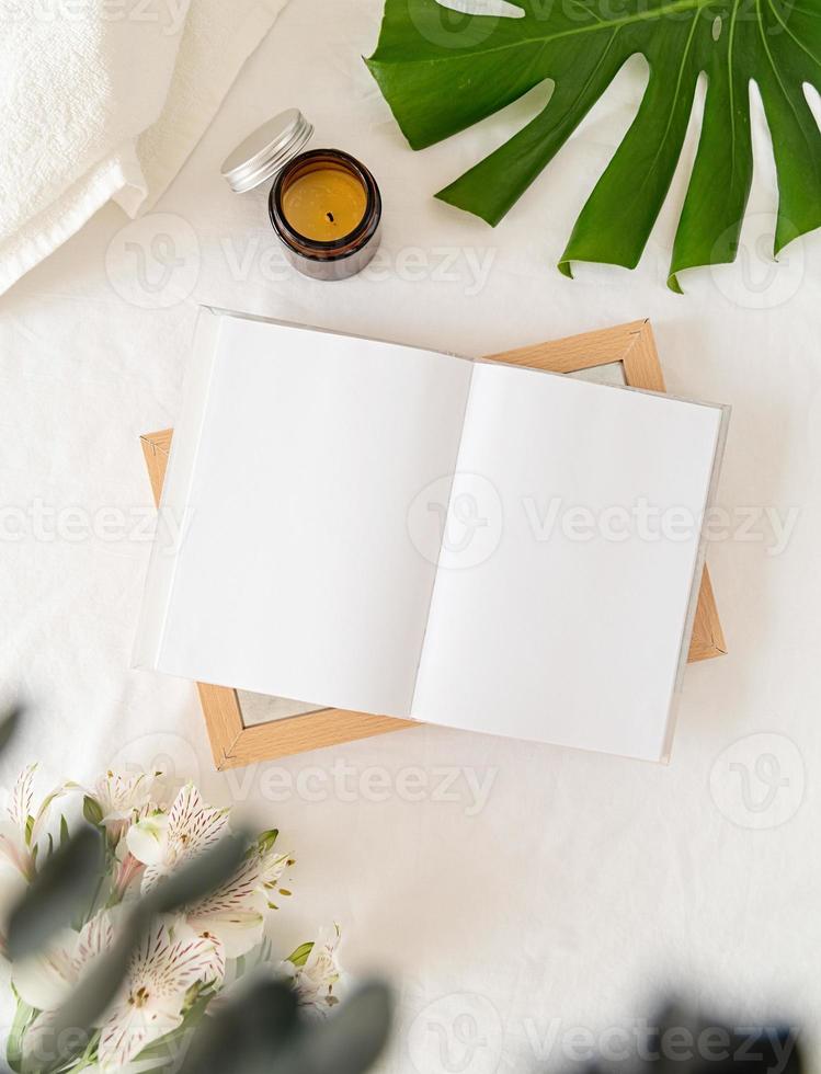 Opened book, candle and flowers top view on white bed. Mock up design photo