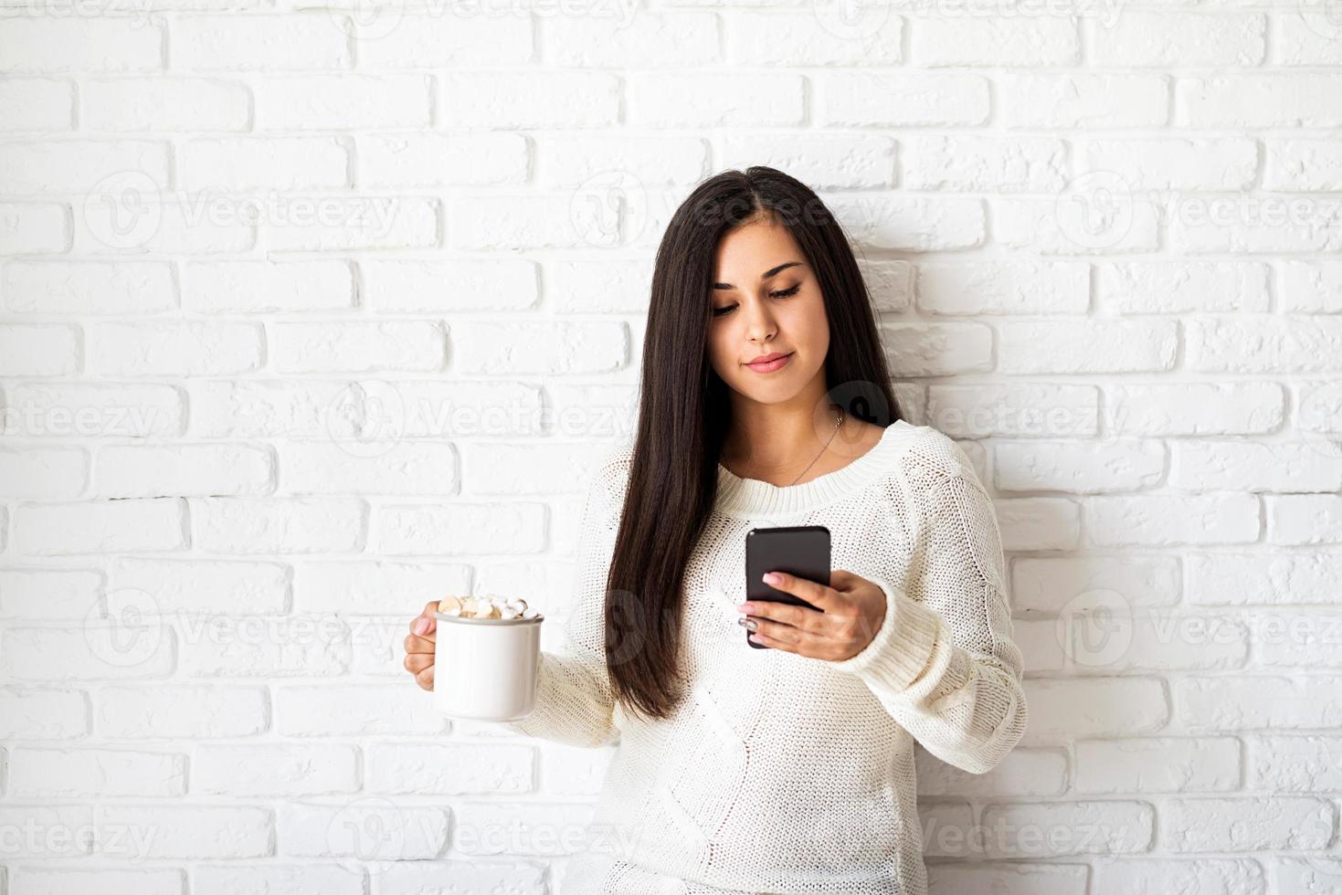 mujer sosteniendo una taza de chocolate con malvavisco y su teléfono foto