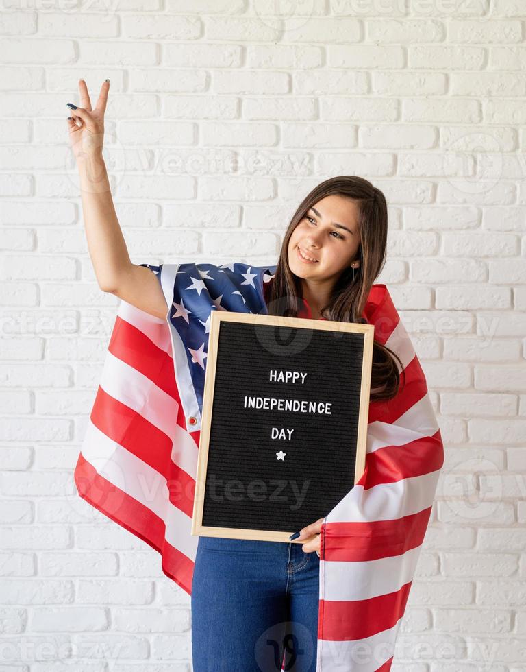 Mujer con bandera americana sosteniendo un tablero de letras con palabras feliz día de la independencia y mostrando el signo de la paz foto